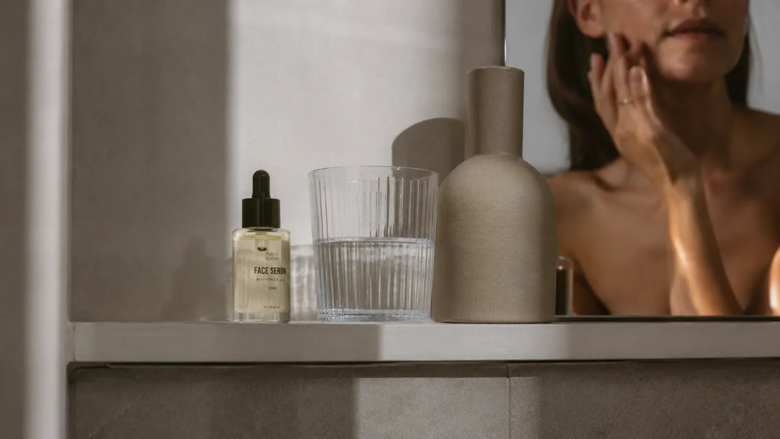 Small amber serum bottle next to a glass of water on a white bathroom shelf with soft morning light