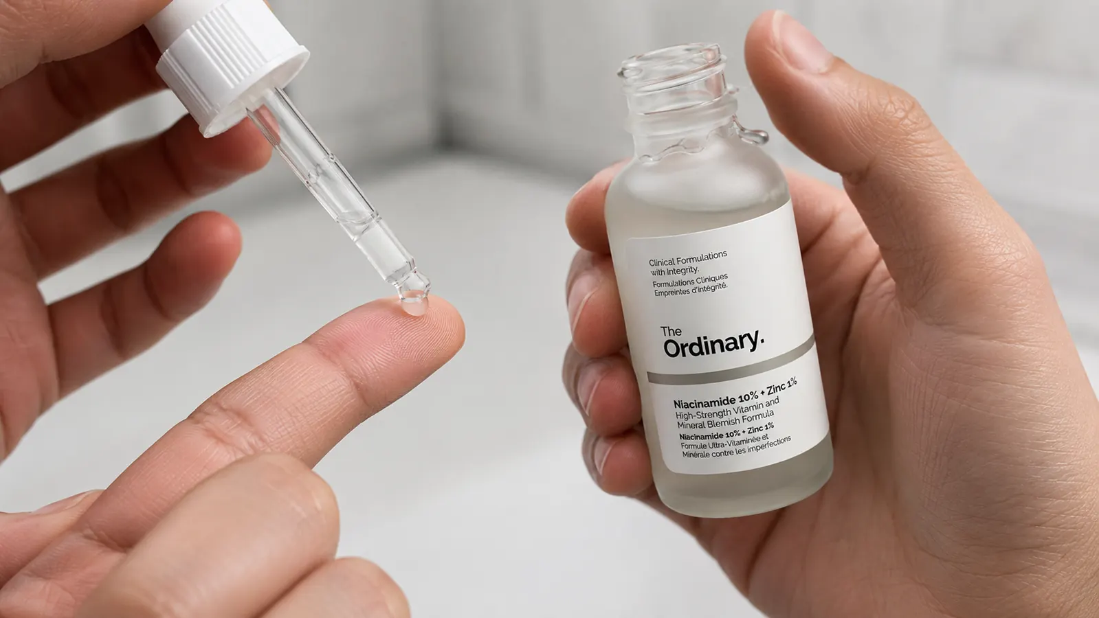 A hand pressing a dropper of clear niacinamide serum onto fingertips above an open palm