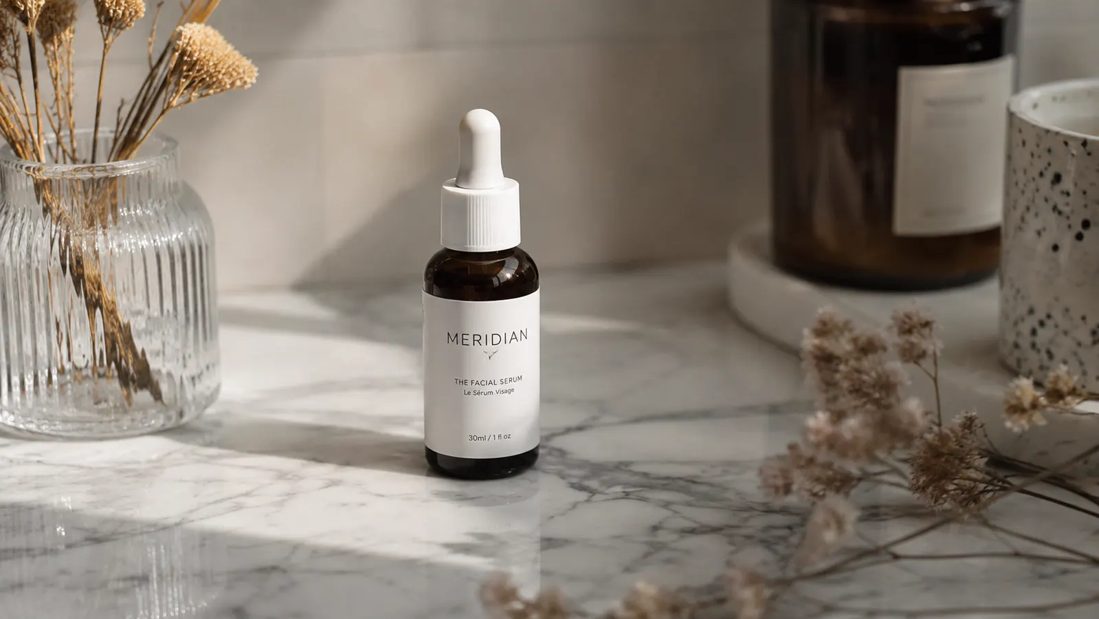 Small brown glass bottle of niacinamide serum on a white marble surface next to a sprig of dried botanicals in soft morning light