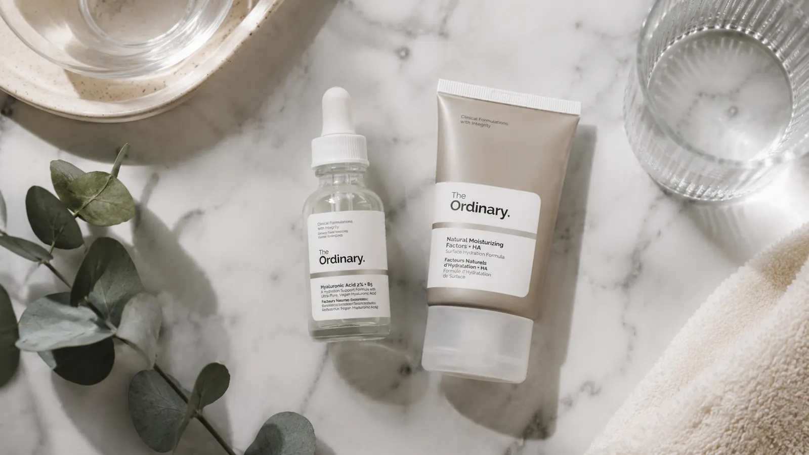 Flatlay of a dropper serum bottle alongside a tube of moisturizer and a small glass of water on a white marble surface