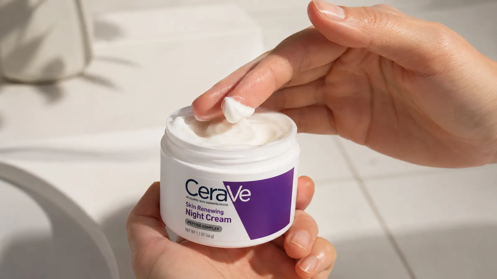 Hand pressing a small amount of white night cream from a jar onto fingertips