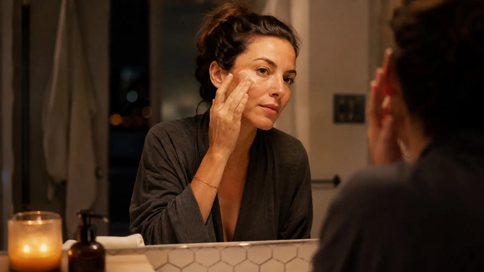 Woman at bathroom vanity in soft evening light applying moisturizer to her face