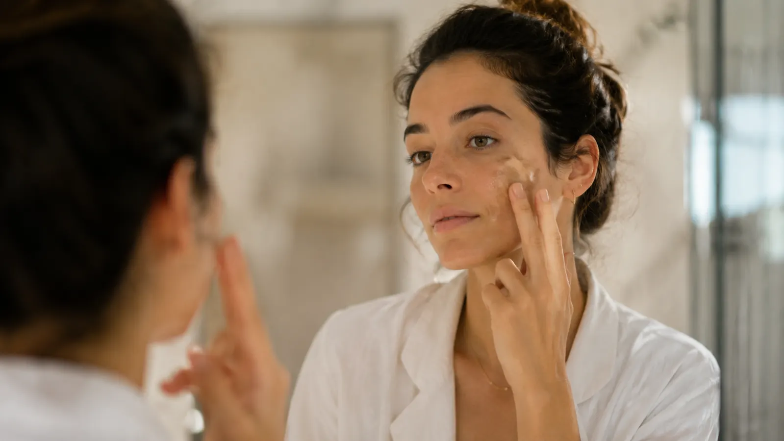 Woman applying tinted sunscreen to her face in warm morning bathroom light, skin looking even and calm