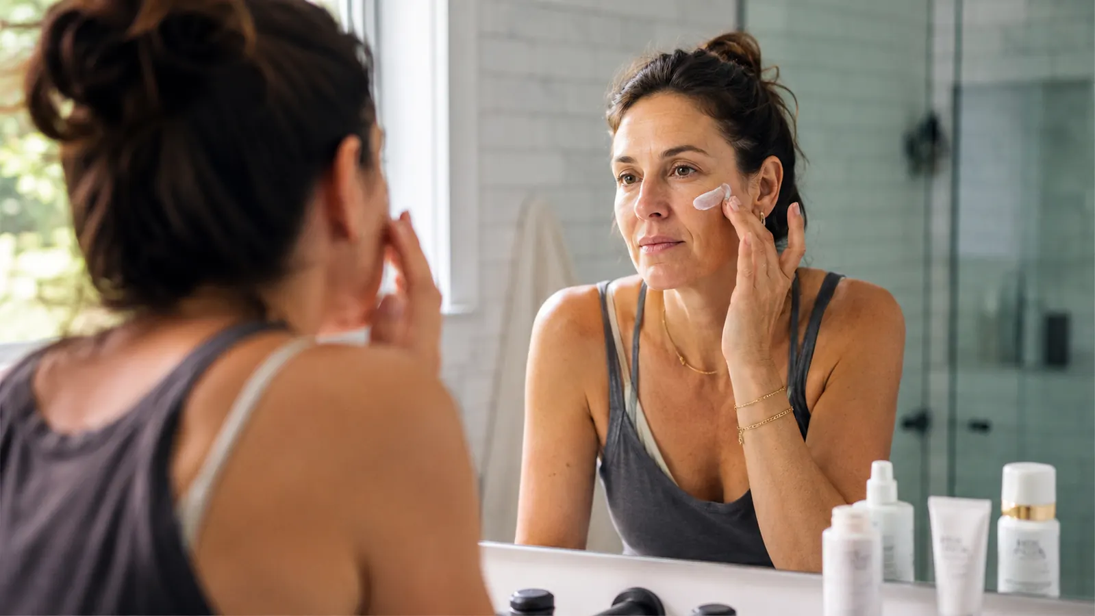 Woman applying sunscreen in the morning mirror, sunlit bathroom, skincare bottles on counter