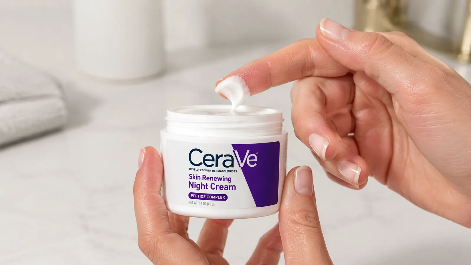 Hands applying a small amount of CeraVe Skin Renewing Night Cream from a white jar onto a fingertip, bathroom mirror visible in background
