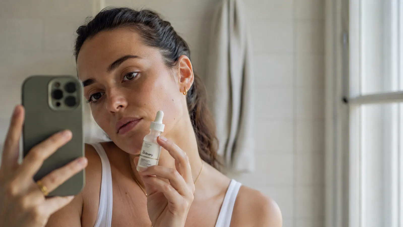A small dropper bottle of niacinamide serum resting on a bathroom shelf beside a cleanser and a light moisturizer, soft morning light coming through a frosted window