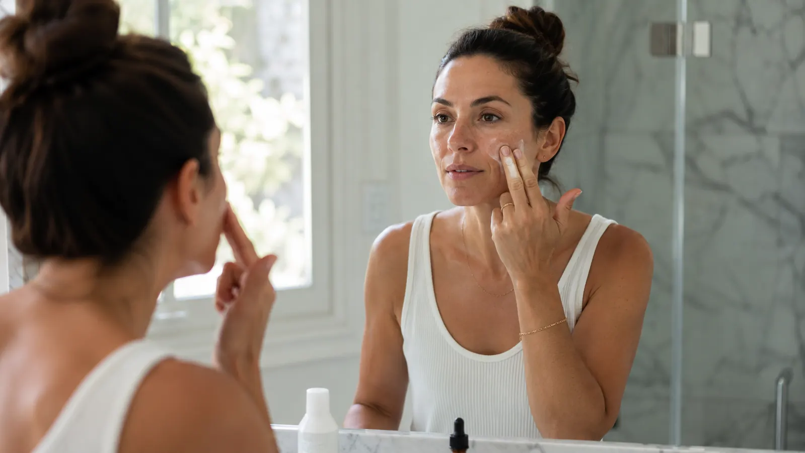 Woman pressing tinted mineral sunscreen into skin at a bathroom mirror in soft morning light