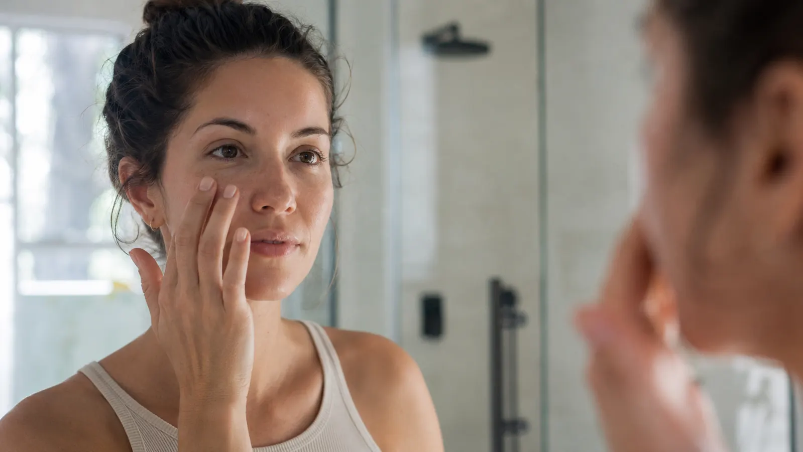 Woman with combination skin applying a clear serum to her cheek in a well-lit bathroom mirror